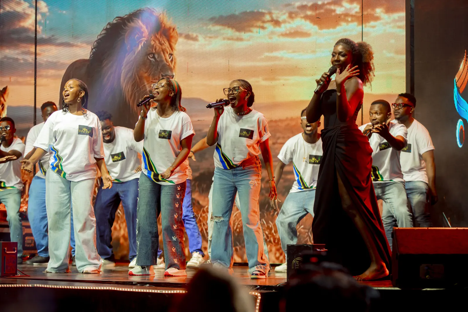 Backing singers in Miss Tanzania T-shirts performing behind a lead vocalist, against a screen showing a lion at sunset